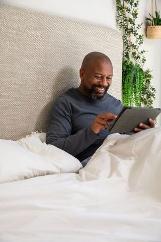 Mature african american man smiling using tablet in bed with hanging plants and calm bedroom