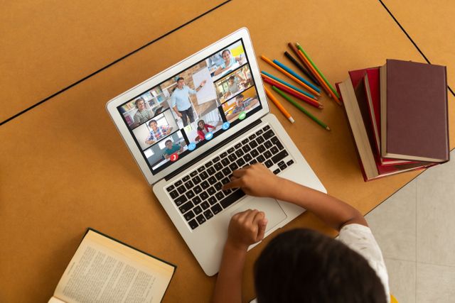 Child sitting at table engaging in online class via laptop, participating in video call with multiple classmates and teacher on screen. Books, colored pencils, and other school supplies scattered on table, emphasizing educational environment. Ideal for representing concepts of distance learning, modern education technology, or pandemic impact on schooling.