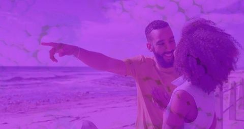 Playful couple pointing toward horizon on seaside boardwalk holding beach ball, purple tint