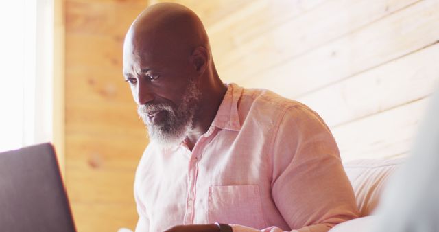 A senior African American man is using his laptop at home in a relaxed, casual setting. Ideal for illustrating themes of remote work, senior technology use, at-home study, and staying connected.