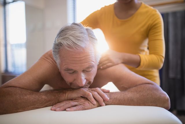 Senior man lying on bed receiving a neck massage from a young female therapist in a hospital ward. Ideal for use in healthcare, wellness, and physical therapy contexts, showcasing professional care and rehabilitation services.