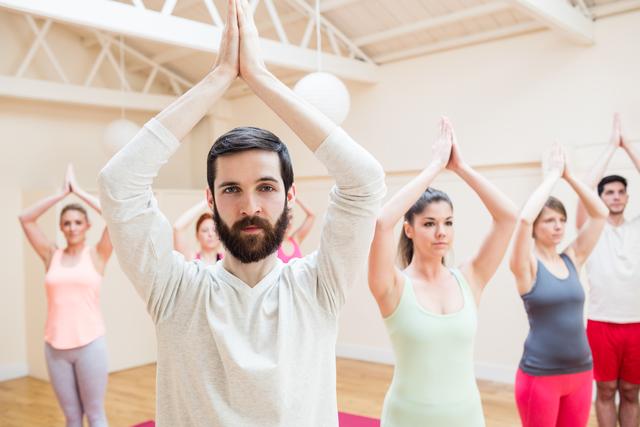 Man practicing tree pose in yoga class with group of people in background. Ideal for promoting fitness, wellness, and mindfulness. Suitable for use in health and fitness blogs, yoga studio advertisements, and wellness programs.