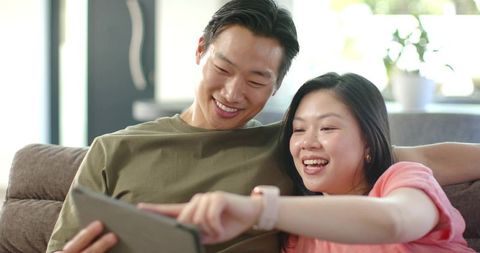 Asian couple relaxing on sofa using tablet together while woman wearing smartwatch