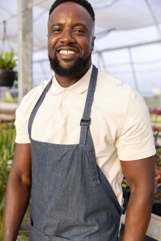 Smiling african american greenhouse grower wearing denim apron among seedlings and plants
