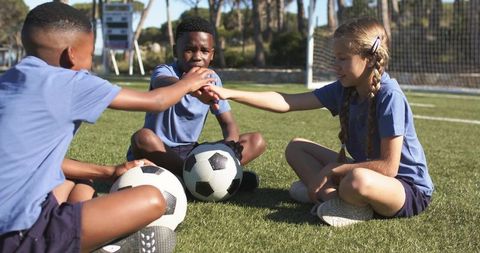 Multiracial youth soccer teammates sitting on grass sharing fist bump with soccer balls