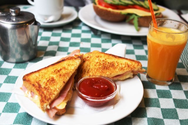 Grilled cheese sandwich with ham on white plate with small ketchup container. Orange juice in clear glass beside sandwich, all placed on green-and-white checkered tablecloth. Ideal for use in restaurant menus, cafe advertisements, and food blog posts emphasizing morning meals.