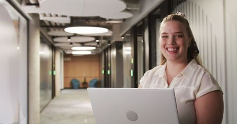 Smiling professional woman holding laptop in modern office corridor