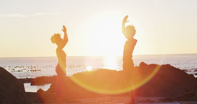 Two people standing on rocky beach, facing each other, arms raised in meditative and stretching pose as sunlight creates a beautiful flare. Ideal for promoting wellness, morning routines, spiritual health, and outdoor activities.
