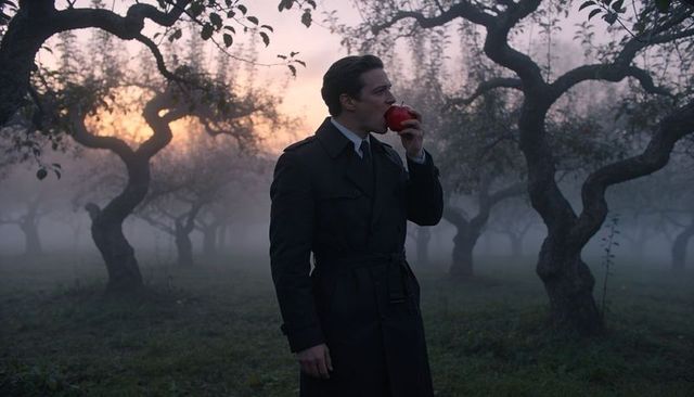 Man in trench coat biting red apple in foggy orchard at sunrise, cinematic backlit mood