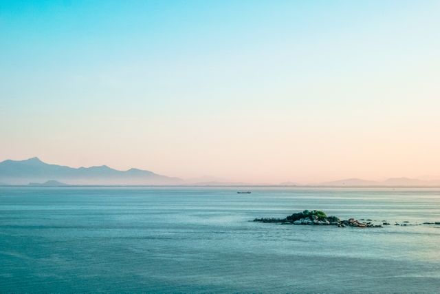 This picturesque scene offers a peaceful ocean view with a small rocky island in the distance, surrounded by calm water and silhouetted mountains on the horizon. Ideal for travel brochures, nature documentaries, relaxation guides, and as a desktop wallpaper to invoke peace and serenity.