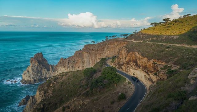 Driving dark suv along winding coastal cliff road with turquoise ocean and rock stacks