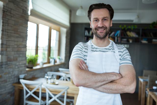 This image depicts a confident bakery owner standing with his arms crossed, smiling warmly. Ideal for use in articles or advertisements about small businesses, entrepreneurship, bakery promotions, or success stories. The cozy and inviting bakery interior adds to the charm, making it suitable for content related to cafes, restaurants, and customer service.