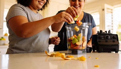 Two women making fruit smoothie at sunlit kitchen island with blender and fresh fruit