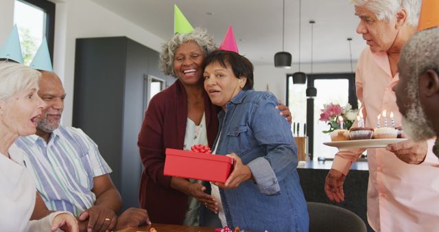 Group of elderly friends celebrating a birthday together. One person is holding a red gift box and others have party hats on. Cake with candles is being brought to the table. This image is ideal for showcasing senior living community events, promoting elderly social activities, or advertising inclusive retirement homes.