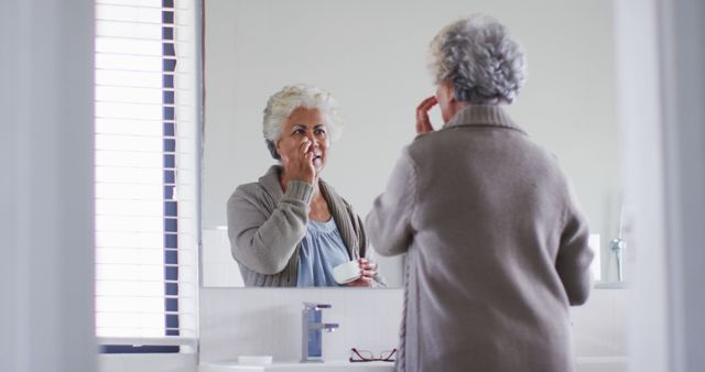 Elderly woman stands in front of a bathroom mirror applying face cream. The scene captures a moment of self-care and a morning skincare routine focused on health and beauty. Ideal for depicting themes of aging gracefully, personal wellness, and daily beauty rituals in publications, advertisements, and health-related articles.