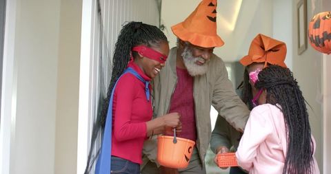 African american grandfather sharing halloween candy with costumed children at doorway