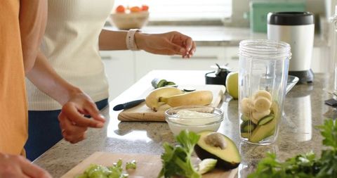 Diverse couple preparing avocado banana green smoothie with yogurt in sunlit kitchen