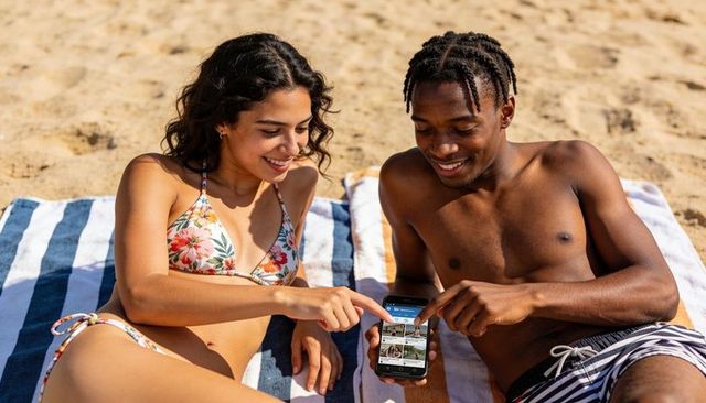 Young couple sharing smartphone while lounging on striped towels at sunny sandy beach