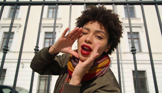 Calling young woman cupping hands by black metal fence wearing olive jacket and striped scarf