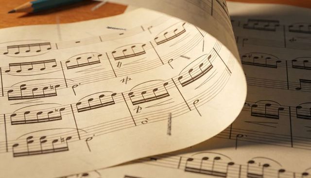 Curled classical sheet music with pencil markings on wooden desk, warm studio light