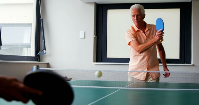 Senior man playing ping pong indoors with friend. Ideal for use in articles about senior activities, healthy lifestyles for older adults, recreational sports, or promoting wellness programs for mature adults. The scene emphasizes active aging, leisure time, and maintaining physical fitness.