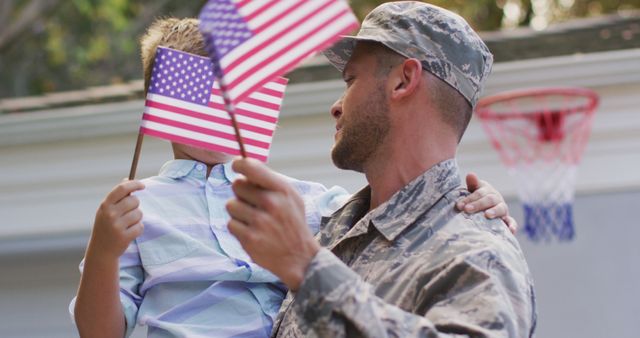 Happy caucasian male soldier carrying his smiling son holding flag in garden outside their house. soldier returning home to family.