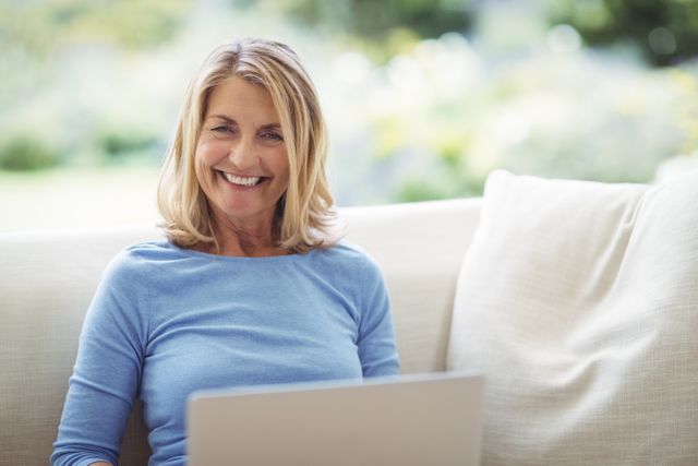 Portrait of smiling senior woman sitting on sofa with laptop in living room at home