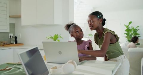 Teen guiding younger girl on laptop at bright modern kitchen table for home learning