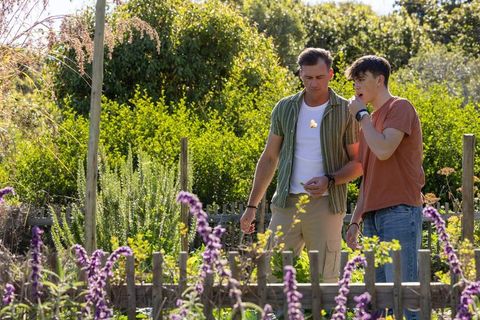 Man mentoring teenage boy in sunny garden planting seeds and inspecting flowers for family bonding