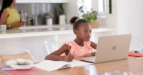 Preteen girl studying on laptop at kitchen table while african mother checking smartphone