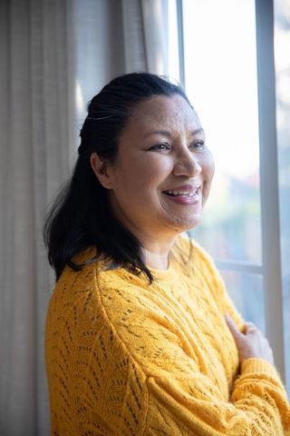 Mature woman smiling and gazing out window in mustard knit sweater, warm serene sunlight