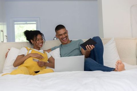 Couple relaxing on bed using laptop and tablet while laughing and holding credit card