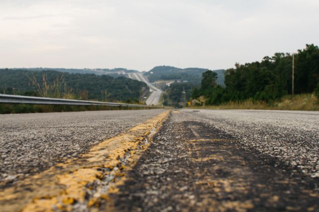 This image shows a close-up view of the road with a focus on the yellow stripe, leading the eye towards a hilly landscape covered in trees and a guardrail on the side. Ideal for themes related to travel, journey, and exploration. Can be used for travel blogs, road trip advertisements, or any content emphasizing nature, adventure, or outdoors.