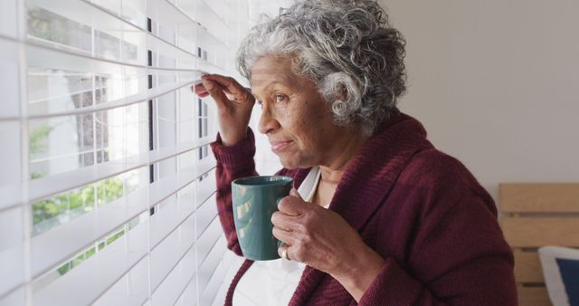 Elderly woman holds green coffee mug and looks out window with a thoughtful expression. She wears a cozy maroon cardigan, suggesting a relaxed morning at home. The white blinds and soft natural light create a calm and serene atmosphere. This image captures everyday moments of contemplation and relaxation. It can be used in articles on aging, mental health awareness, lifestyle blogs, or advertisements for home products and senior services.