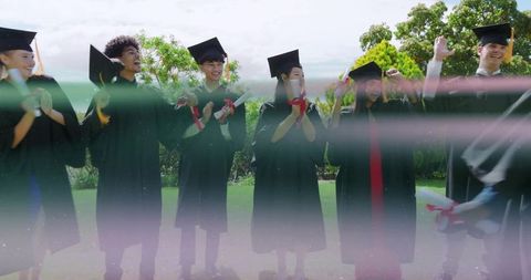 Graduates cheering on campus lawn in caps and gowns holding diplomas, joyful celebration