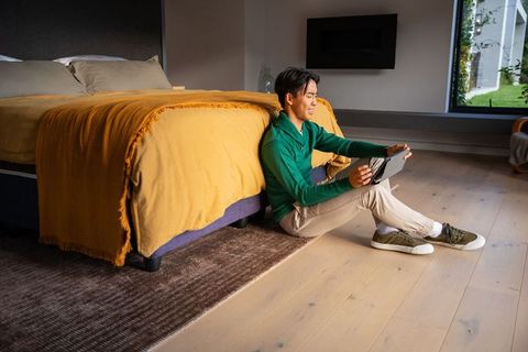 Asian man sitting on bedroom floor using tablet leaning against bed with mustard throw