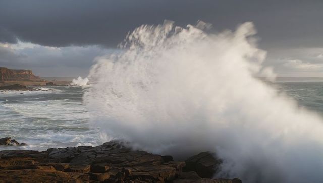 Crashing wave sending towering white spray over rocky coastal cliffs under stormy sky