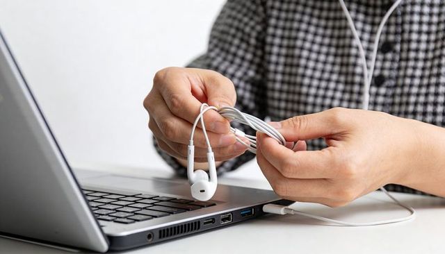 Hands coiling white wired earbuds beside laptop on modern desk closeup cable management