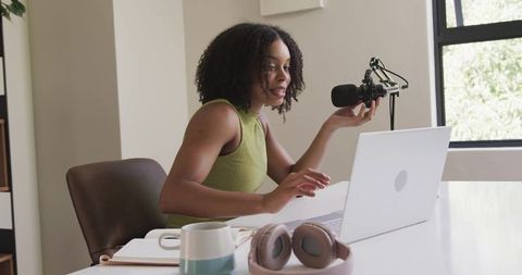 African american woman podcasting at home desk with laptop, microphone, headphones