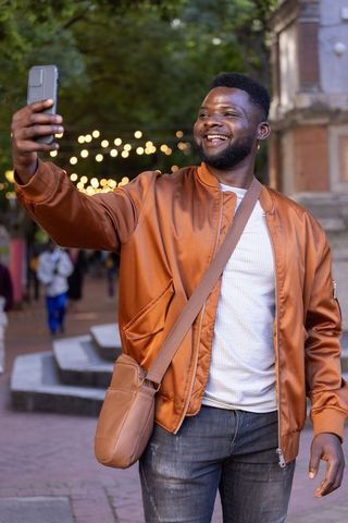 African american man taking selfie at twilight wearing brown bomber jacket and crossbody