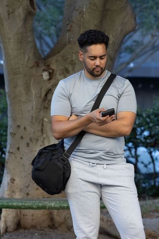 African american man standing in park wearing gray athletic wear holding smartphone and duffel