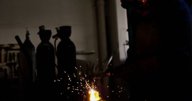 Factory worker utilizing a welding torch, generating bright sparks in a dimly lit industrial workplace. Perfect for illustrating themes of heavy industry, manufacturing processes, blue-collar jobs, industrial safety precautions, or skilled labor. Could be used for educational materials, advertisements in the industrial sector, or articles on modern manufacturing.