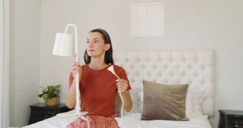 Woman installing lamp in modern bright bedroom on cozy bed