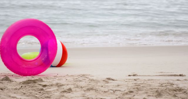 Colorful inflatable beach toys, including a pink ring float and a striped beach ball, are on a sandy shore by the ocean. This scene captures the essence of a fun summer day, perfect for advertisements, travel promotions, and material to evoke sea vacation vibes.