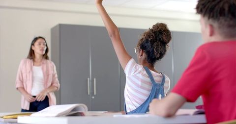 Teenage girl raising hand and participating in classroom lesson with teacher observing