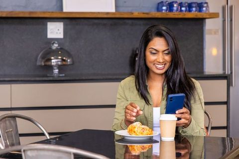 Indian woman smiling while checking smartphone and reaching for croissant in kitchen