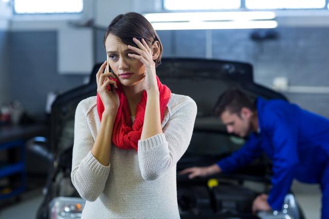 Worried woman talking on mobile phone while mechanic inspects car in background. Useful for illustrating automotive repair services, customer service scenarios, emergency situations, and stress management. Ideal for blogs, articles, and advertisements related to car maintenance, repair shops, and customer support.