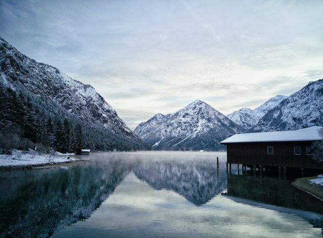 Snow covered mountains reflecting in clear, calm lake water with a small wooden cabin on the shore. Ideal for use in travel advertisements, nature magazines, website background images, relaxation or meditation content promoting outdoor retreats or scenic getaways.