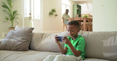 African american father working while son playing mobile game on couch in bright open-plan home