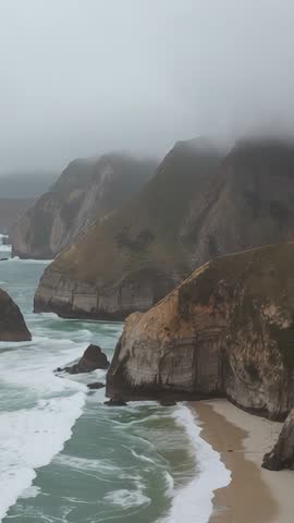Vertical aerial video drifting along misty sea cliffs with pounding waves and empty beach